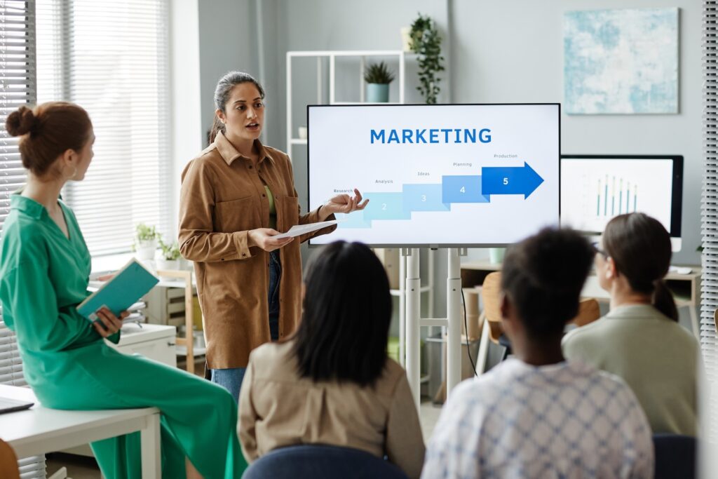 Una mujer presenta un diagrama de marketing en una pantalla ante cuatro personas en una oficina con ventanas, estanterías y plantas.