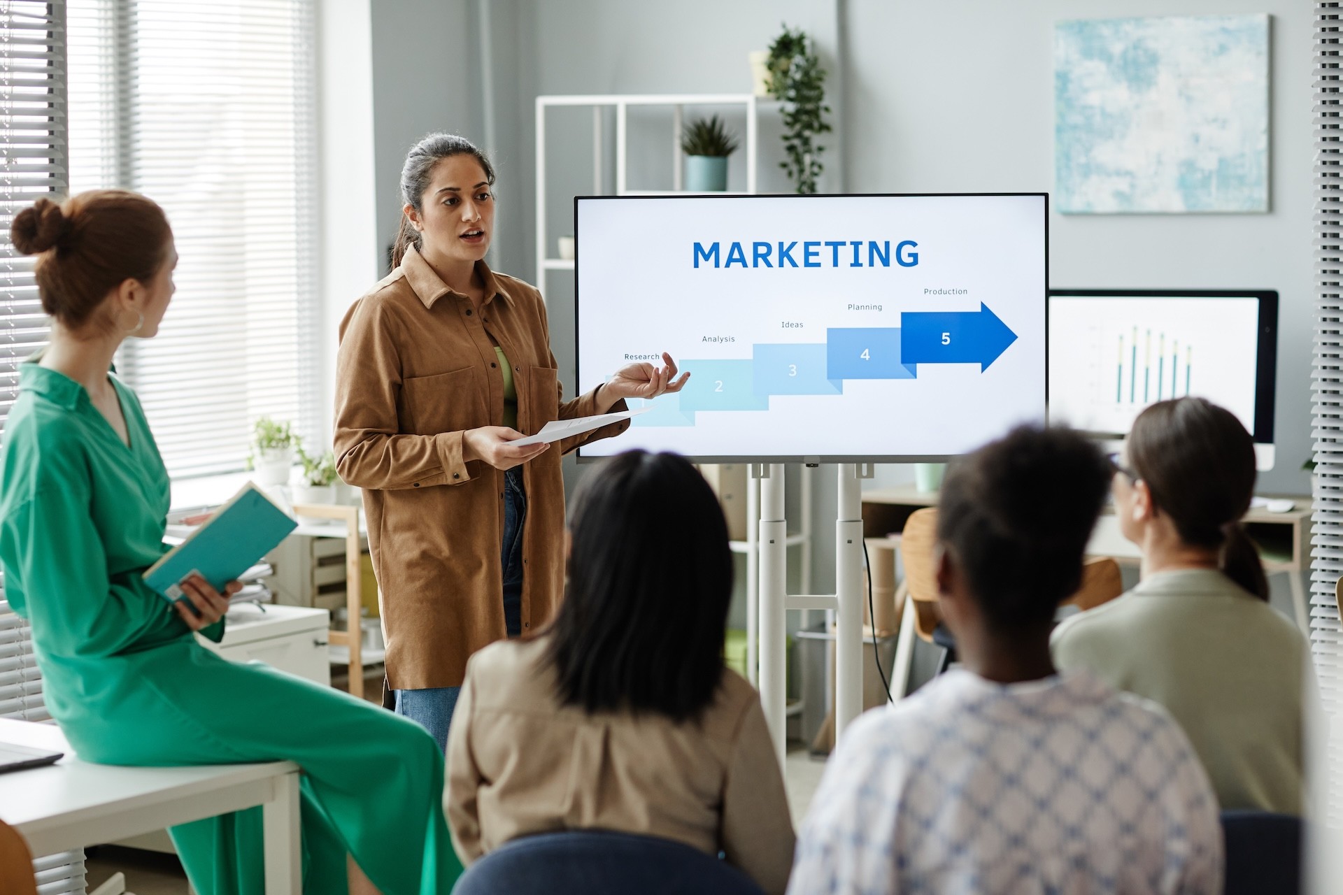 Una mujer presenta un diagrama de marketing en una pantalla ante cuatro personas en una oficina con ventanas, estanterías y plantas.